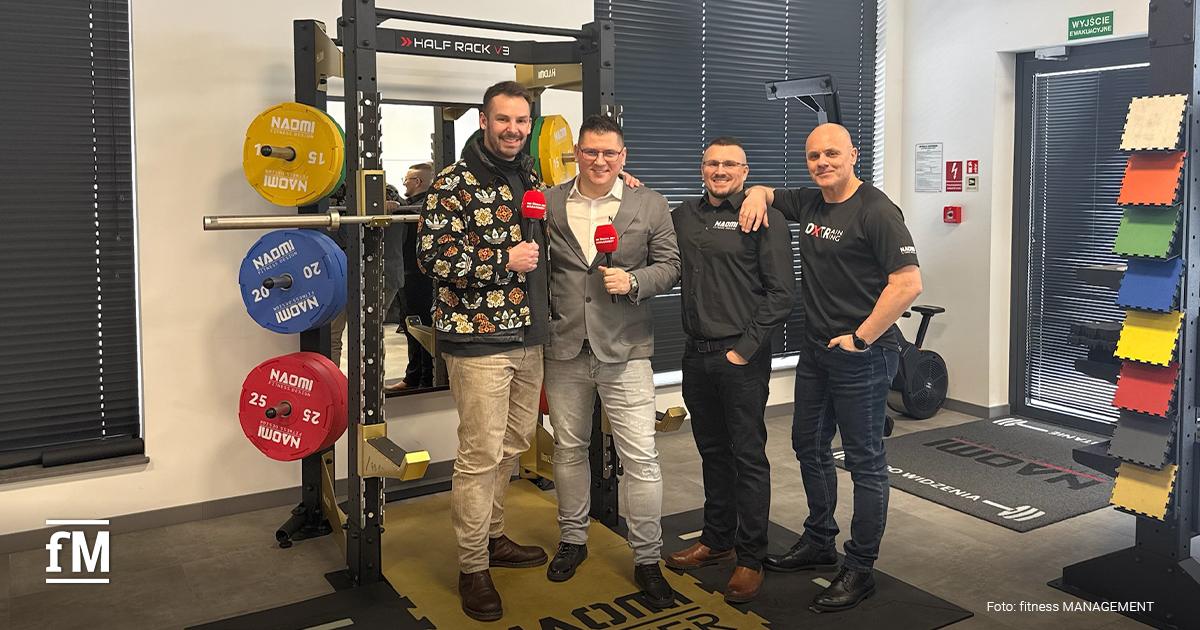 Vier Männer (Janosch Marx, Marek Brzózka, Robert Kolanko und Marek Prusiński) stehen neben einem Half Rack mit farbigen Hantelscheiben, schwarzer Trainingsboden mit gelben Markierungen und Fenster mit Jalousien.