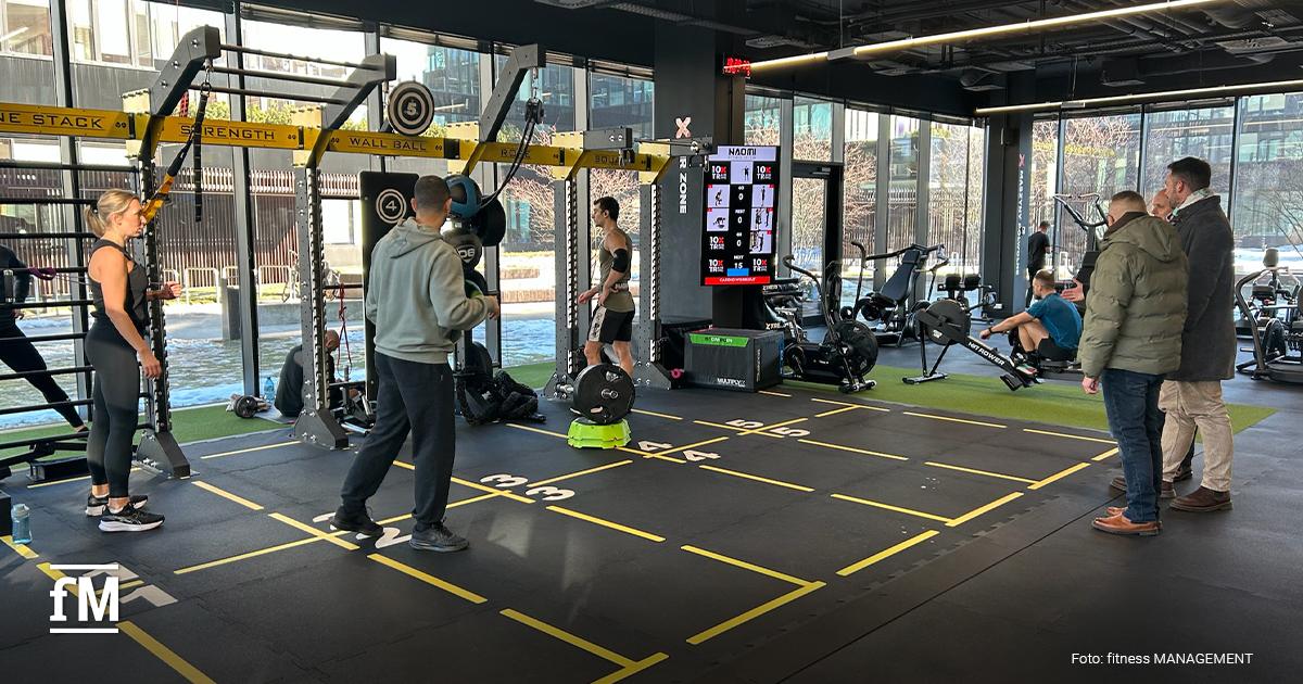 Fitnessstudio mit gelb markierten Trainingsfeldern am Boden, Functionalrig entlang großer Fenster, mehrere trainierende Personen und rechts ein digitaler Trainingsscreen.