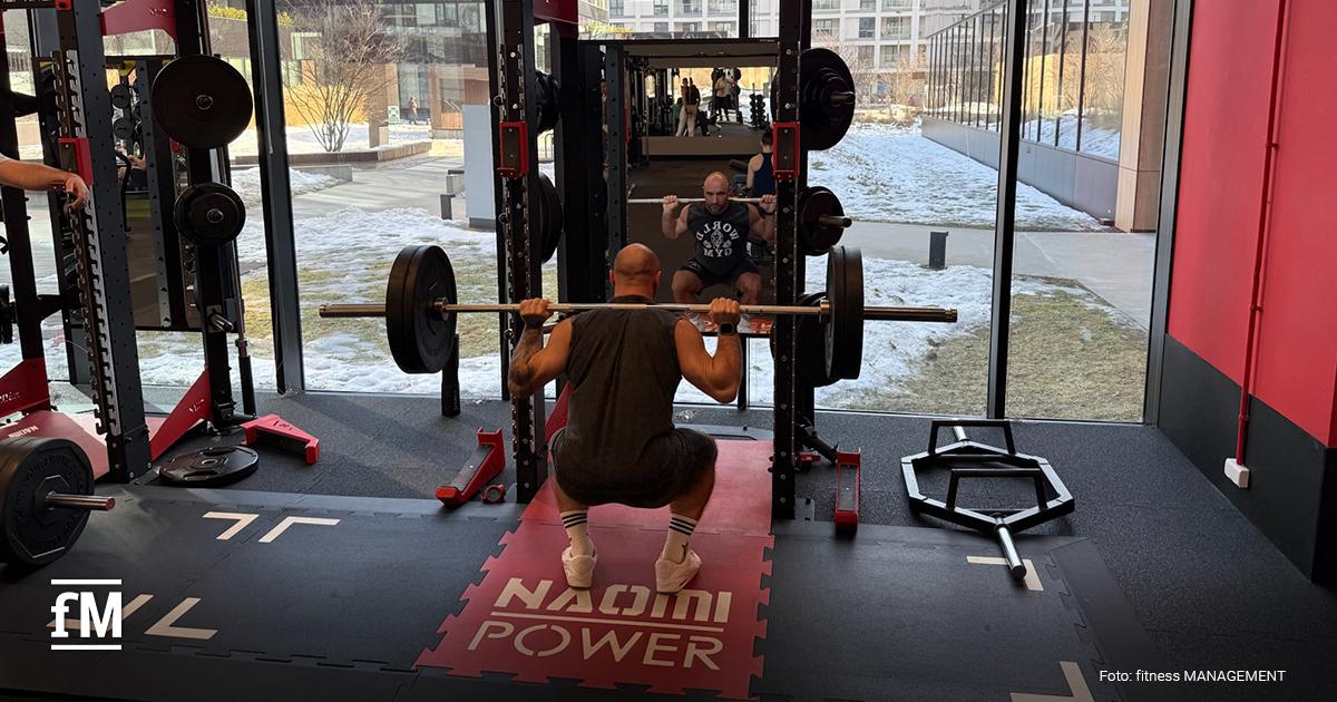 Half Rack vor Fensterfront, ein Mann führt eine Langhantelkniebeuge aus, Spiegel reflektiert die Bewegung, rechts liegt eine Hexbar auf dem Boden.