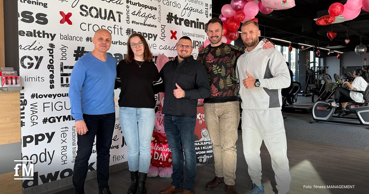 Mehrere Personen stehen nebeneinander in einem Fitnessstudio vor einer Wand mit Trainingsbegriffen, im Hintergrund Cardiogeräte und rote Ballondekoration.