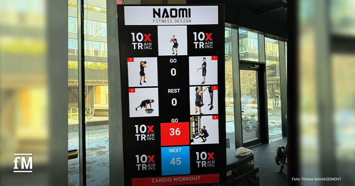 Großer vertikaler Bildschirm mit 10XTraining-Logo, Übungsdarstellungen und Countdownanzeige, positioniert vor einer Glasfront.