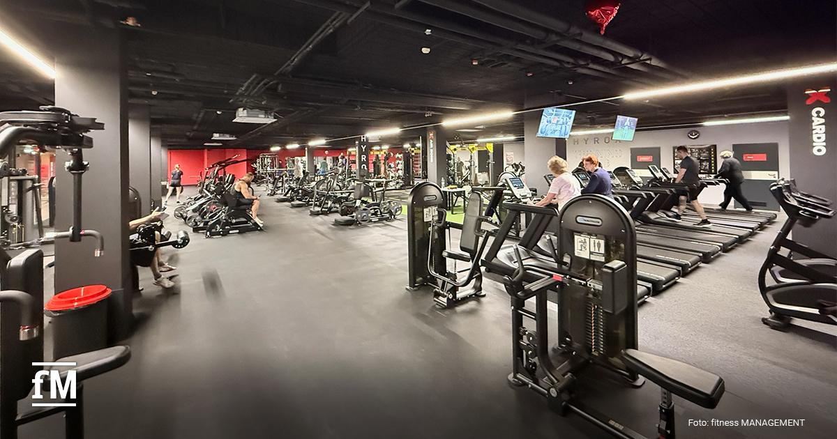 Großer Fitnessraum mit mehreren Laufbändern in Reihenaufstellung, trainierende Personen auf Cardiogeräten, dunkle Decke mit Lichtleisten.