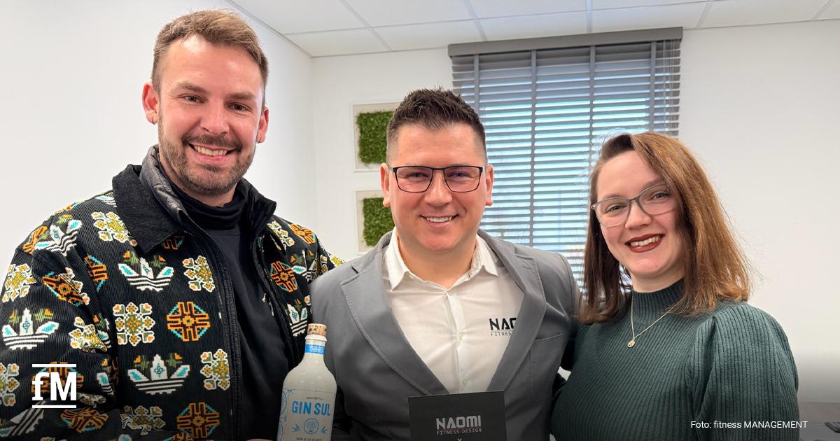 Drei Personen (Janosch Marx, Marek Brzózka und Alisha Dittmer) stehen in einem Büro, eine Person hält eine Flasche, im Hintergrund Fenster mit Jalousien.