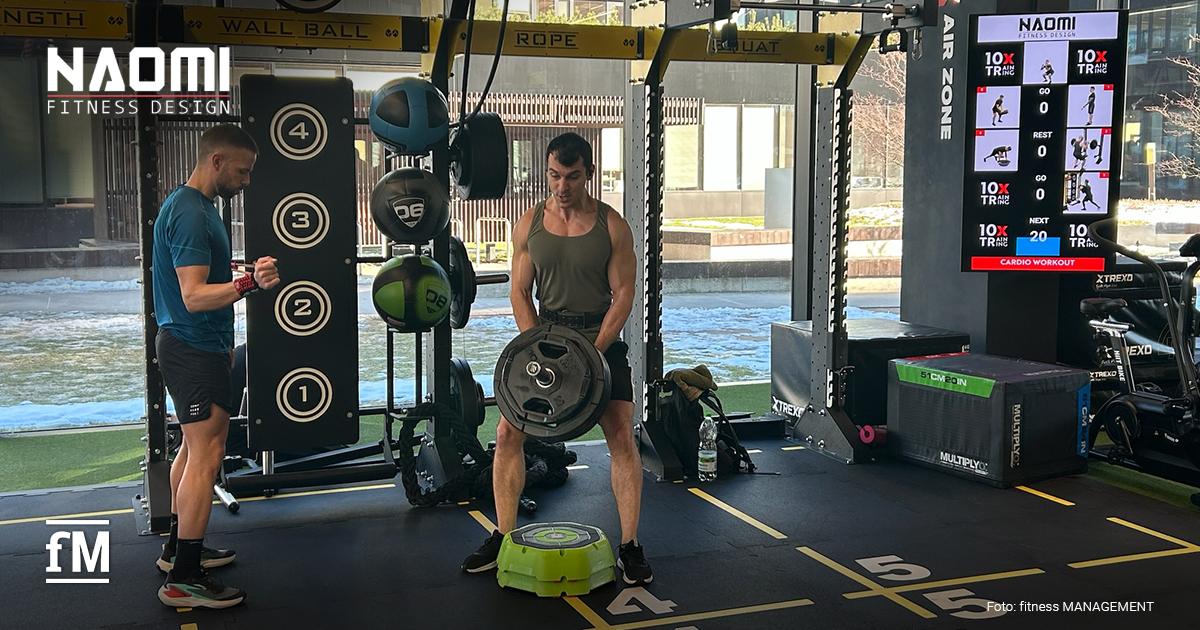 NAOMI FITNESS DESIGN Polen Functional Rig mit Medizinbällen und nummerierter Zielwand, zwei Männer trainieren mit Gewichten auf markierten Bodenfeldern, rechts ein vertikaler 10XTraining-Bildschirm.