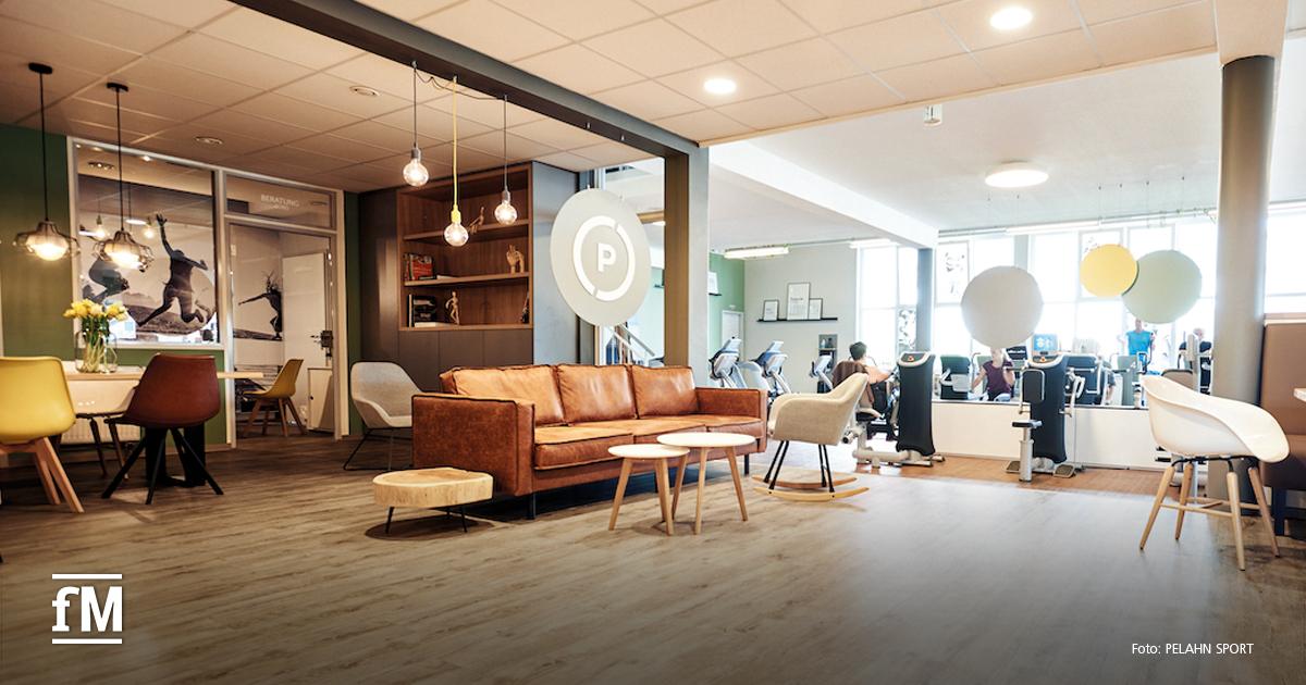 Offener Innenraum mit Sofa, Sesseln und Tischen, angrenzend eine Trainingsfläche mit Fitnessgeräten und trainierenden Personen.