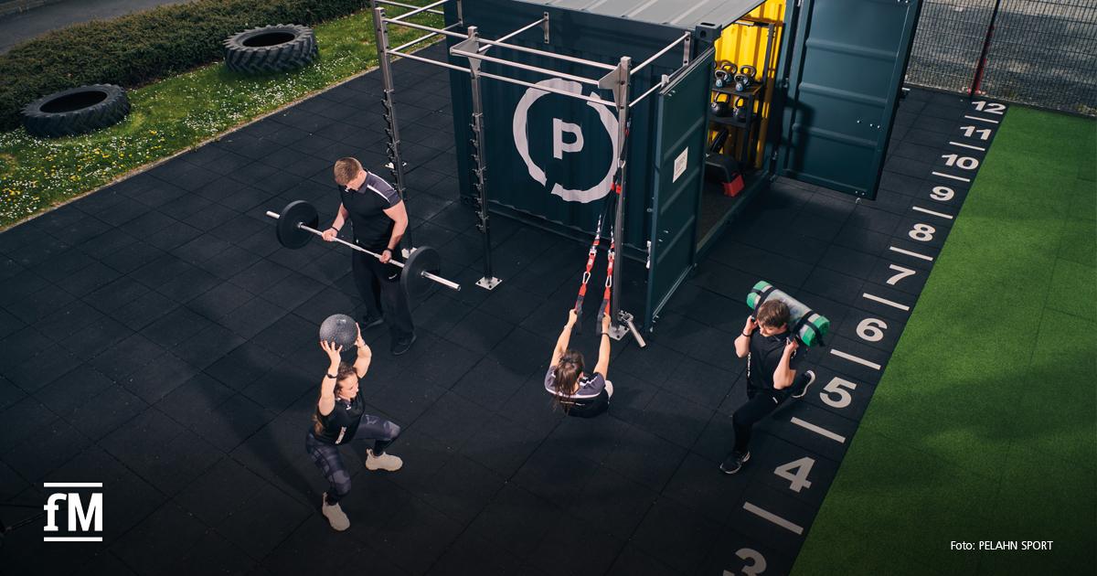 Vier Personen trainieren im Außenbereich auf schwarzem Boden an einem Container mit Logo, mit Langhantel, Medizinball, Schlingentrainer und Gewichtssack.