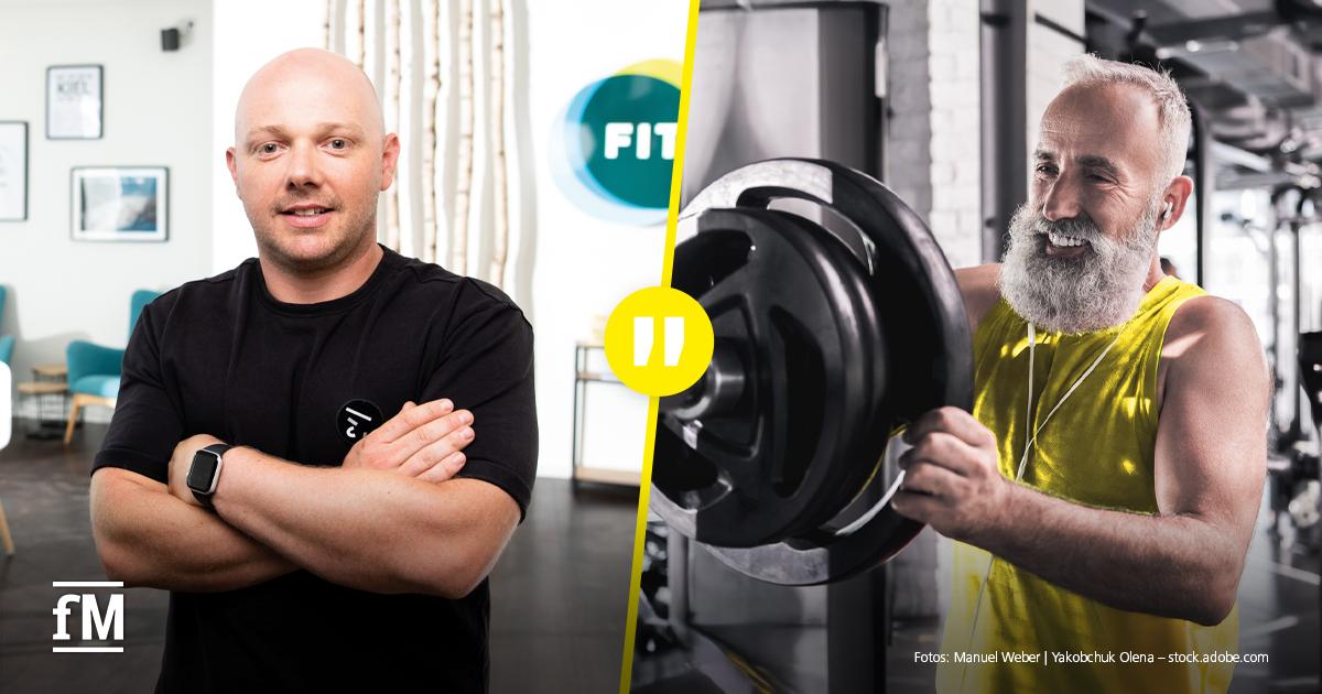 Performance is Health Porträt eines Mannes mit Brille und verschränkten Armen vor dunklem Hintergrund; trägt ein helles Sweatshirt mit UFC GYM Schriftzug