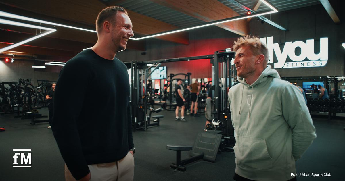 Zwei Männer stehen auf der Trainingsfläche eines Studios und unterhalten sich zwischen Fitnessgeräten