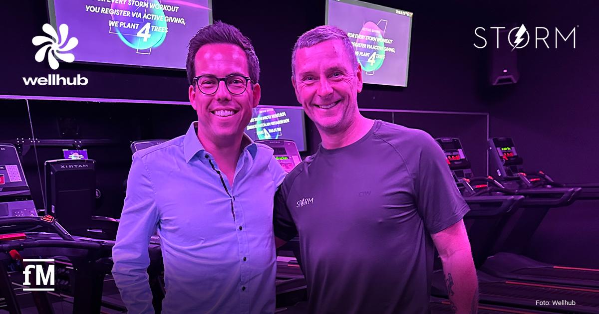 STORM x Wellhub Zwei Personen stehen in einem STORM Fitnessstudio vor Laufbändern, im Hintergrund sind Bildschirme mit Trainingsinformationen zu sehen. Oben links ist das Wellhub-Logo, oben rechts das von STORM und unten links das fM-Signet, alles in Weiß.