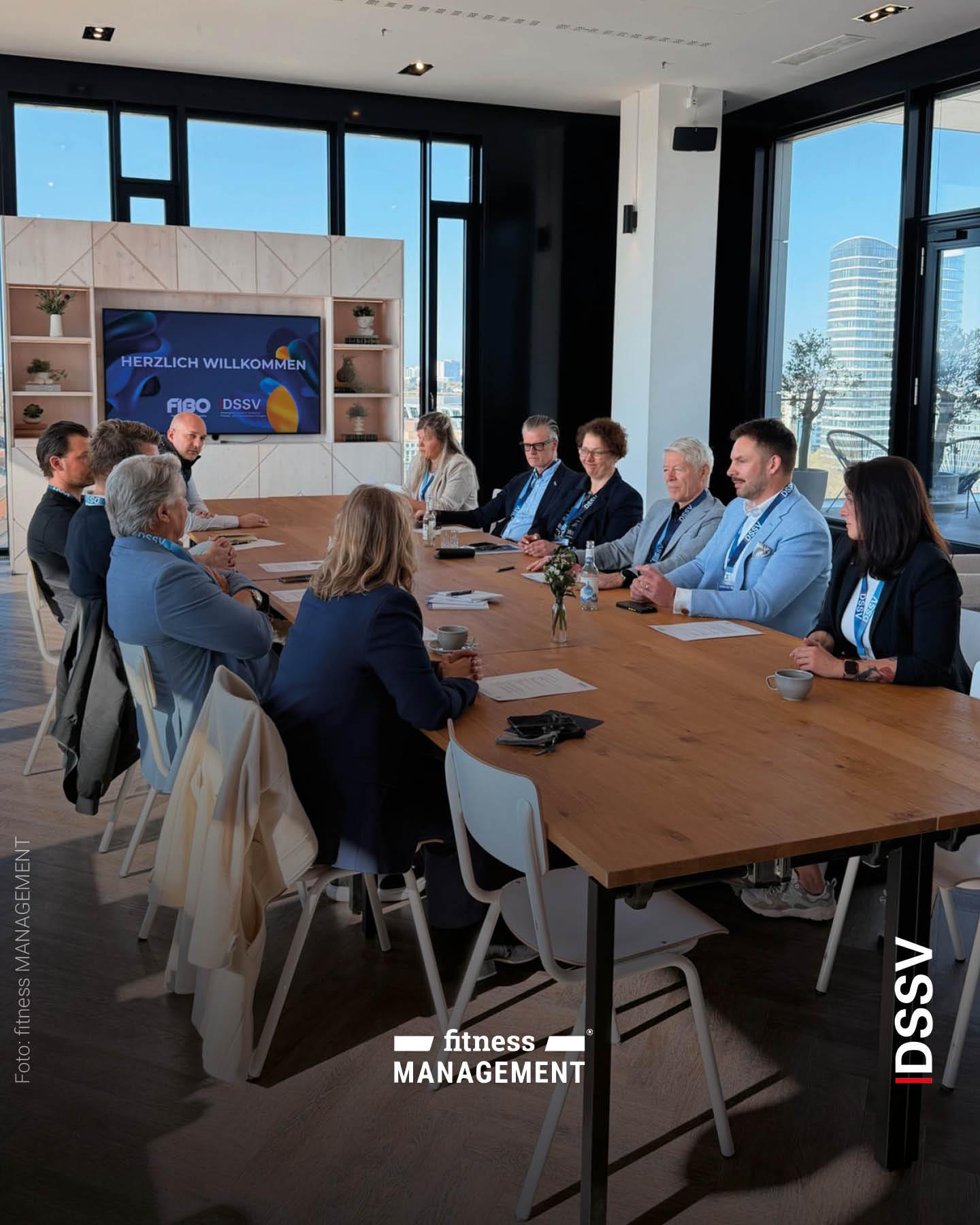 ehrere Personen sitzen an einem großen Tisch in einem modernen Konferenzraum und nehmen an einem Meeting teil, im Hintergrund ein Bildschirm mit Begrüßungsfolie