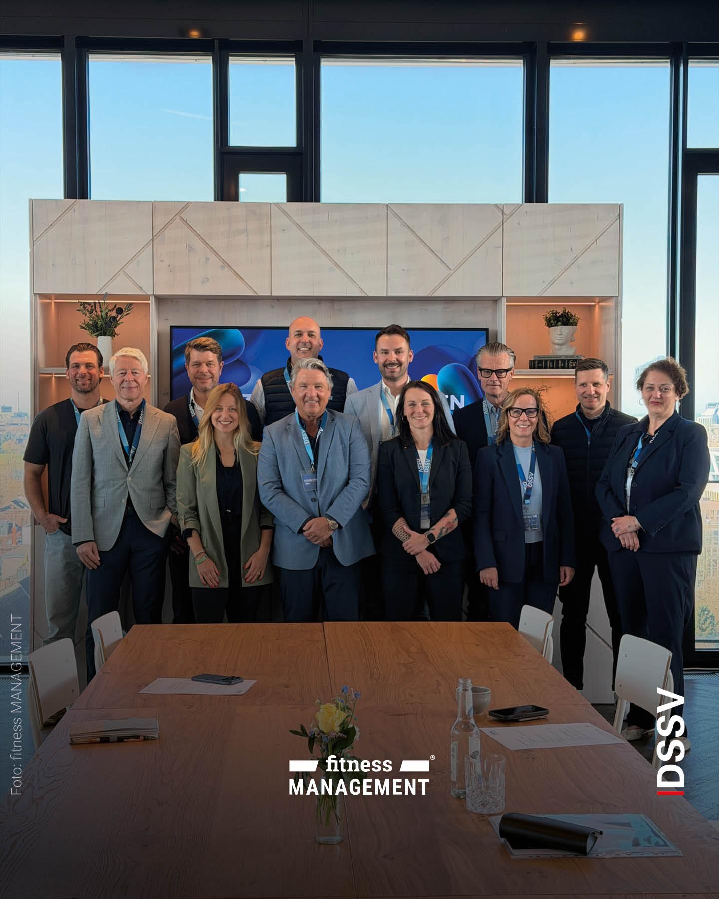 Gruppe von Teilnehmern steht hinter einem Konferenztisch und posiert gemeinsam für ein Gruppenfoto in einem hellen Raum mit großen Fenstern