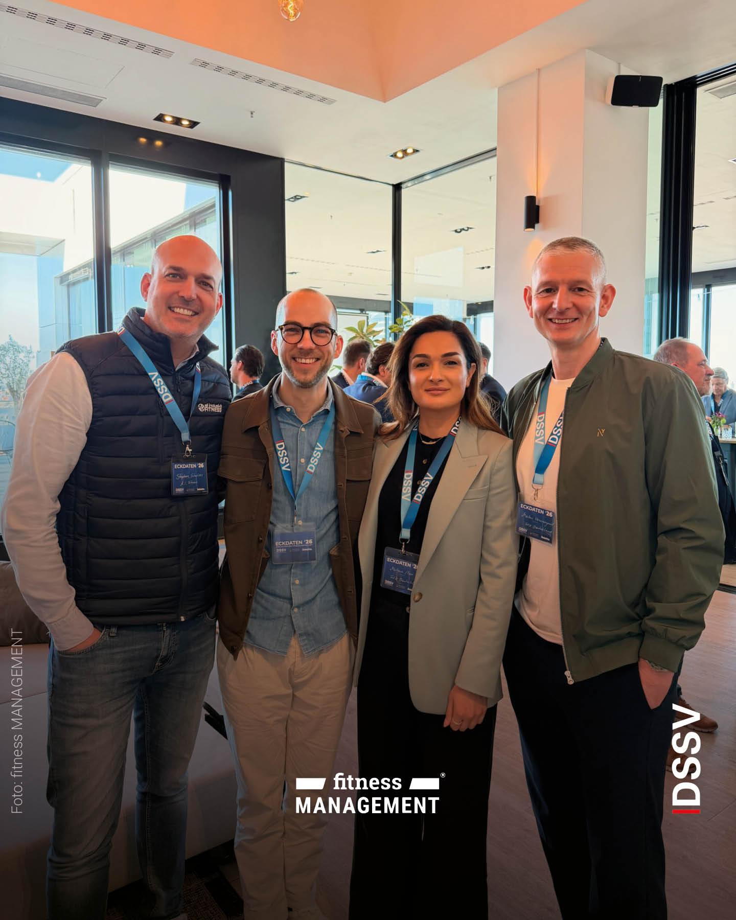 Vier Personen stehen eng zusammen in einem Innenraum und schauen lächelnd in die Kamera, alle tragen Namensschilder der Veranstaltung