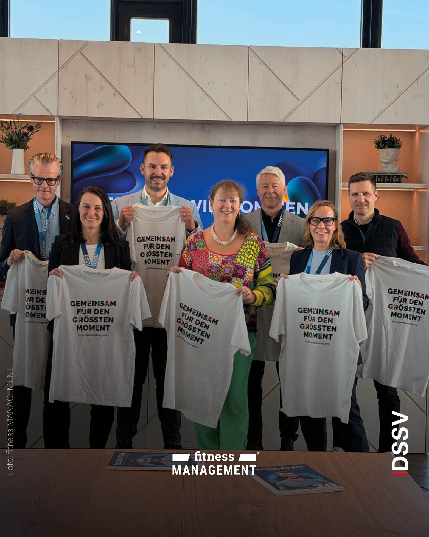 Eine Gruppe von Personen hält weiße T-Shirts mit dem Aufdruck „Gemeinsam für den größten Moment“ vor sich und posiert für ein Foto