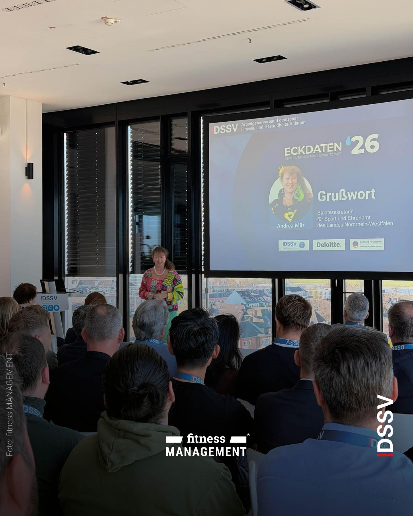 Eine Frau steht vor einem Publikum und hält einen Vortrag, hinter ihr ist eine Präsentationsfolie mit dem Titel „Eckdaten ’26“ zu sehen