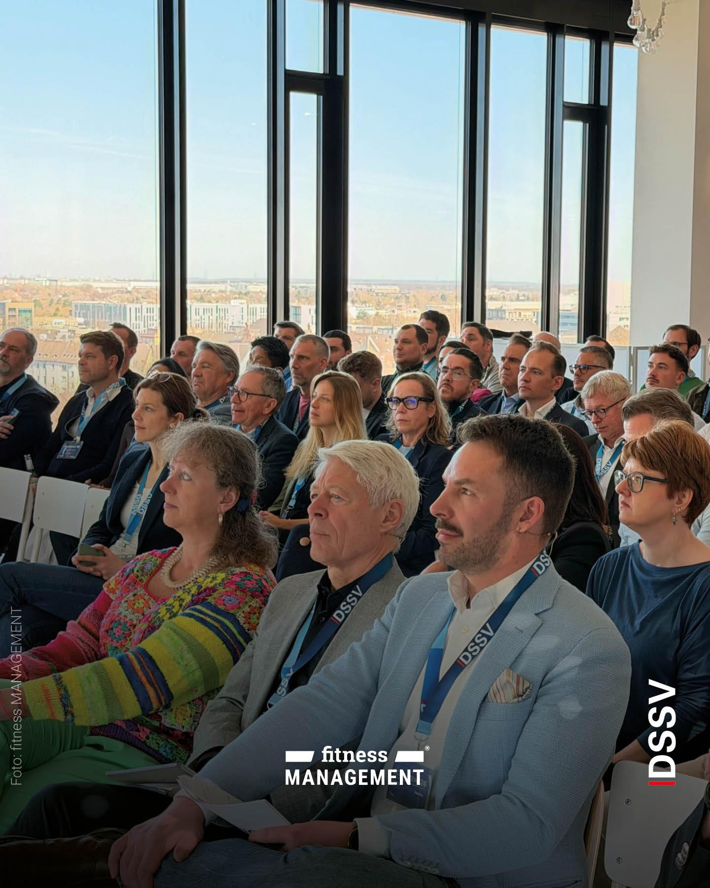 Publikum sitzt in Reihen und hört aufmerksam einem Vortrag zu, im Vordergrund mehrere Personen mit Namensbändern in einem hellen Raum