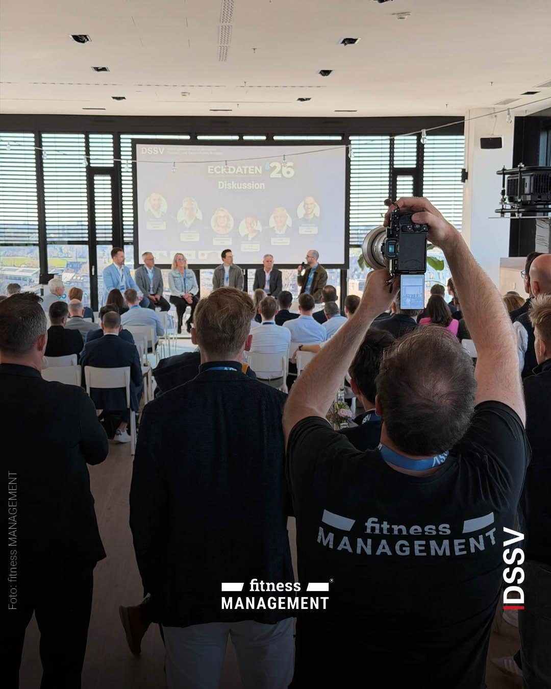 Ein Fotograf hält eine Kamera hoch und fotografiert eine Podiumsdiskussion, während im Hintergrund mehrere Personen auf einer Bühne sitzen und ein Publikum zuhört