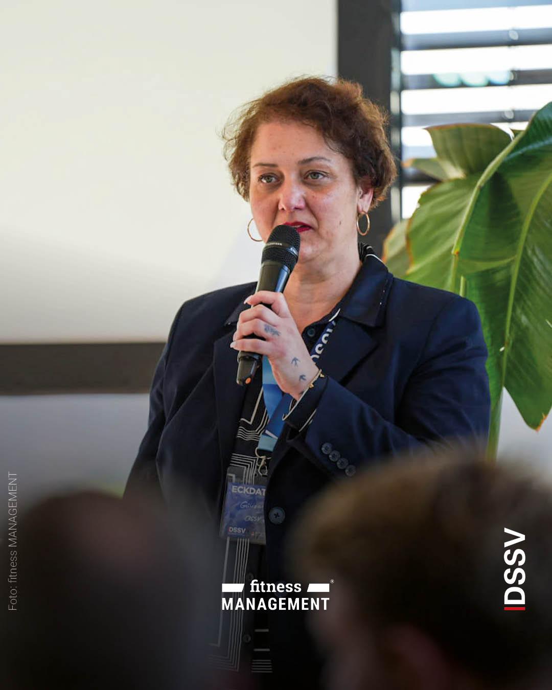 Eine Frau steht vor Publikum und spricht in ein Mikrofon, neben ihr ist eine große Zimmerpflanze zu sehen