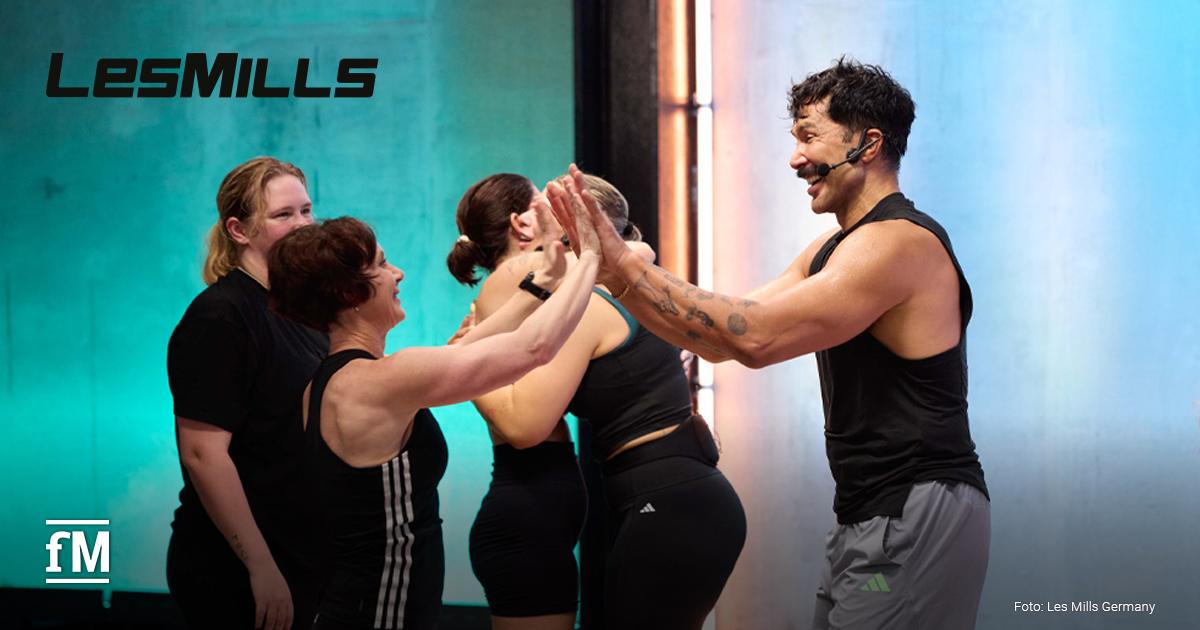 Vordergrund rechts: Trainer mit Headset und schwarzem ärmellosen Shirt steht seitlich zur Kamera und gibt zwei Frauen ein High Five. Links und mittig stehen drei Teilnehmerinnen in Sportkleidung in einer Gruppenfitnessumgebung. Hintergrund zeigt farbig beleuchtete Studiowand.