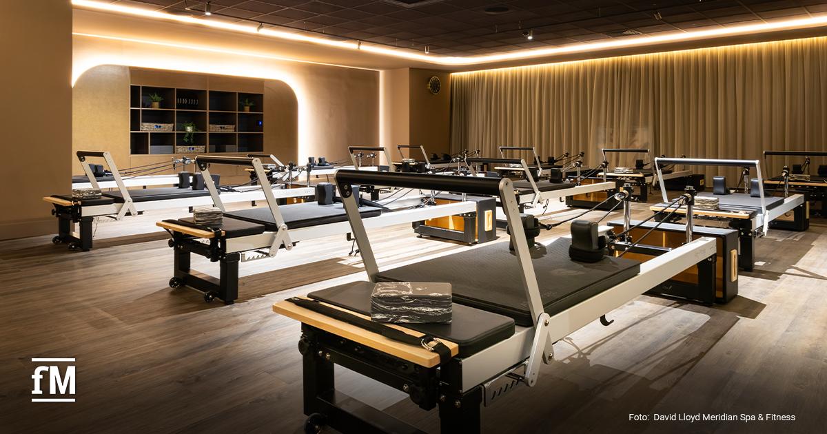 Raum mit mehreren Reformer-Pilates-Geräten, Holzfußboden, Regalen und indirekter Deckenbeleuchtung.