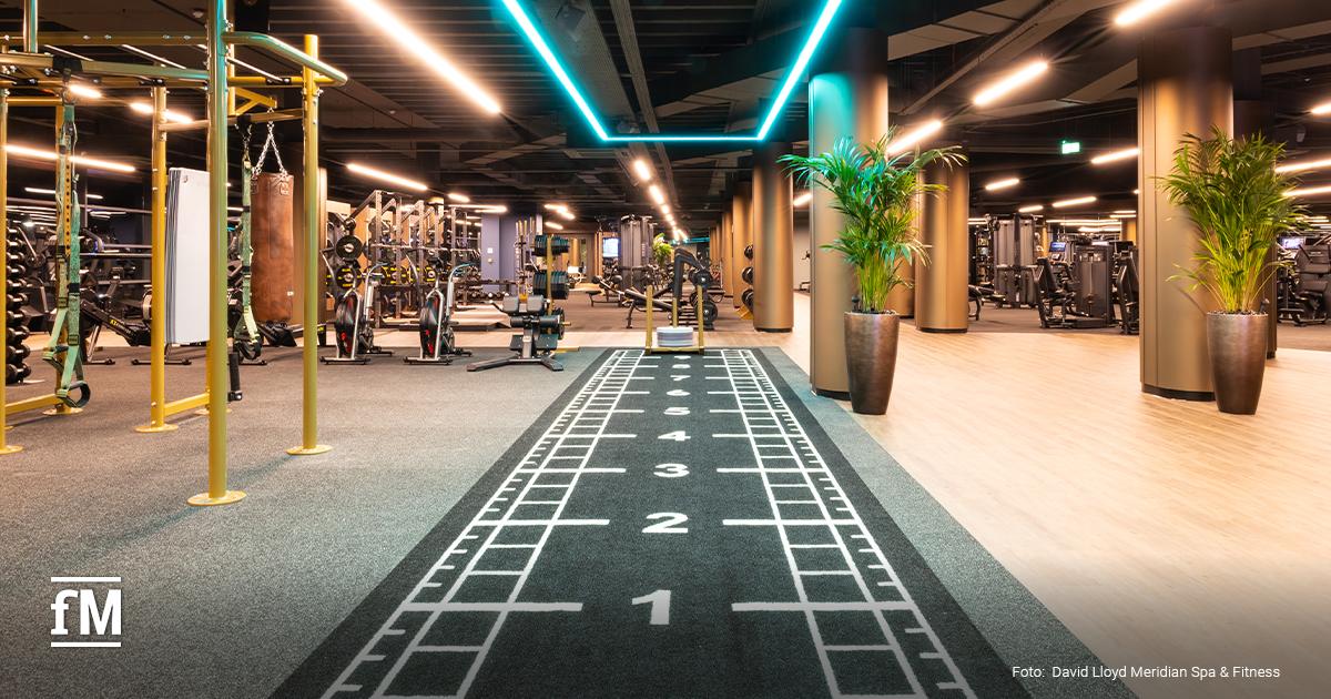 Großer Fitnessraum mit Laufbahnmarkierung auf dem Boden, Kraftgeräten, Säulen und LED-Deckenbeleuchtung.