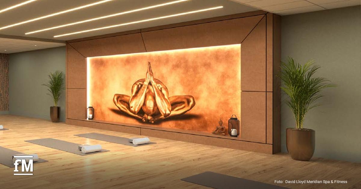 Raum mit Yogamatten auf Holzfußboden vor großer beleuchteter Wandgrafik einer meditierenden Figur, Pflanzen und Laternen an den Seiten.