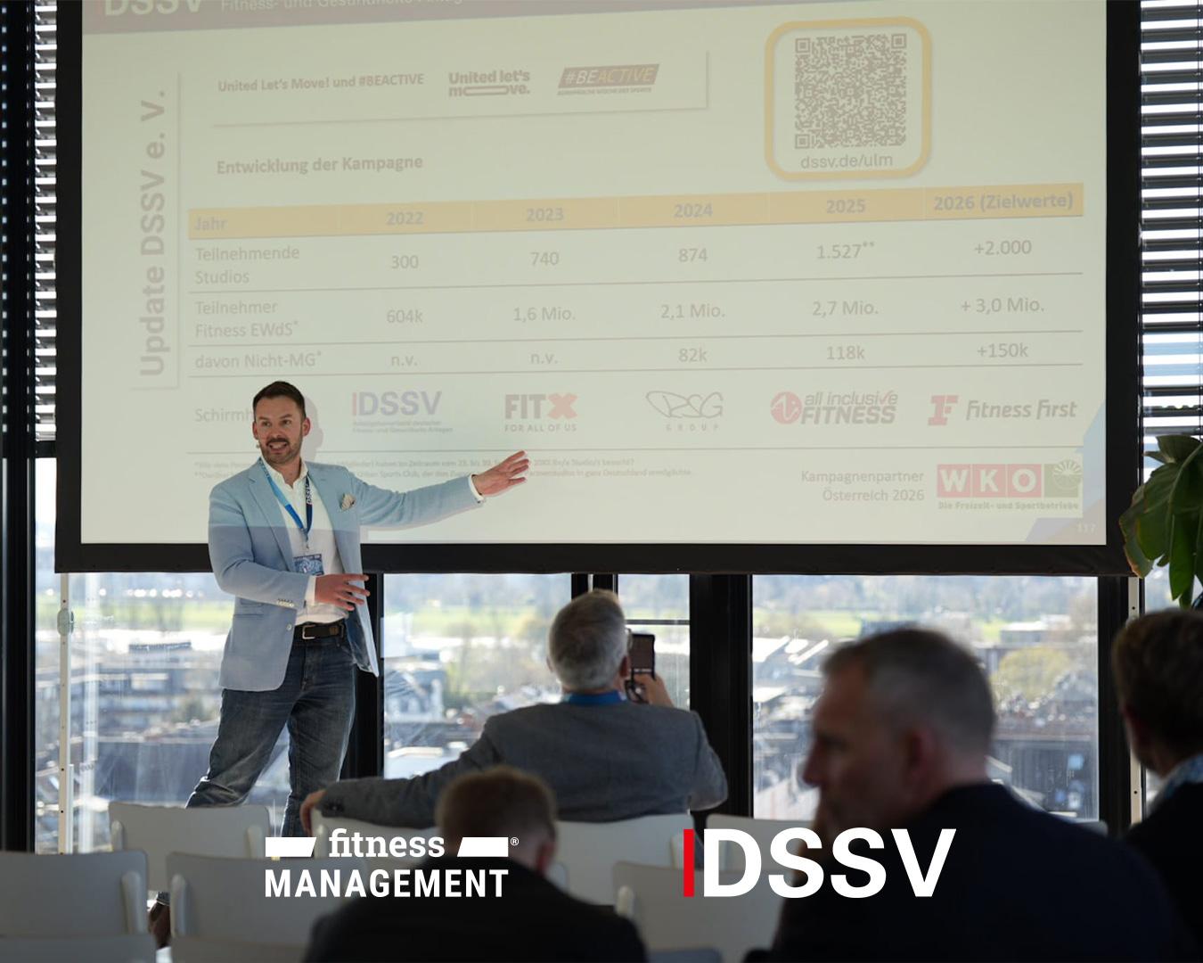 Ein Referent steht vor einer großen Präsentationsfolie und zeigt auf eine Grafik, während das Publikum vor ihm sitzt