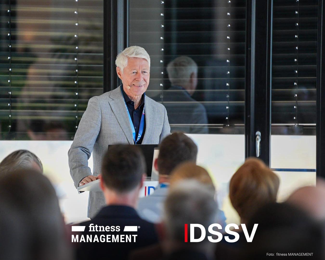 Ein älterer Mann steht hinter einem Rednerpult und spricht zu einem Publikum. Vor ihm sitzen Zuhörer, während hinter ihm Fenster mit reflektierenden Oberflächen zu sehen sind