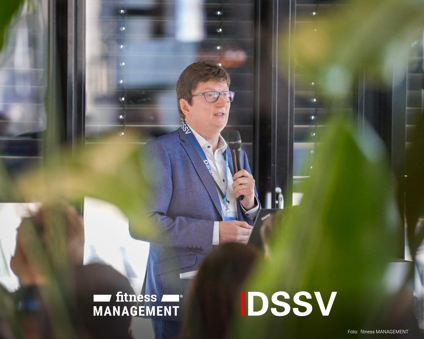 Ein Mann mit Brille hält ein Mikrofon in der Hand und spricht vor einem Publikum. Im Vordergrund sind unscharfe Pflanzen und Zuhörer zu erkennen