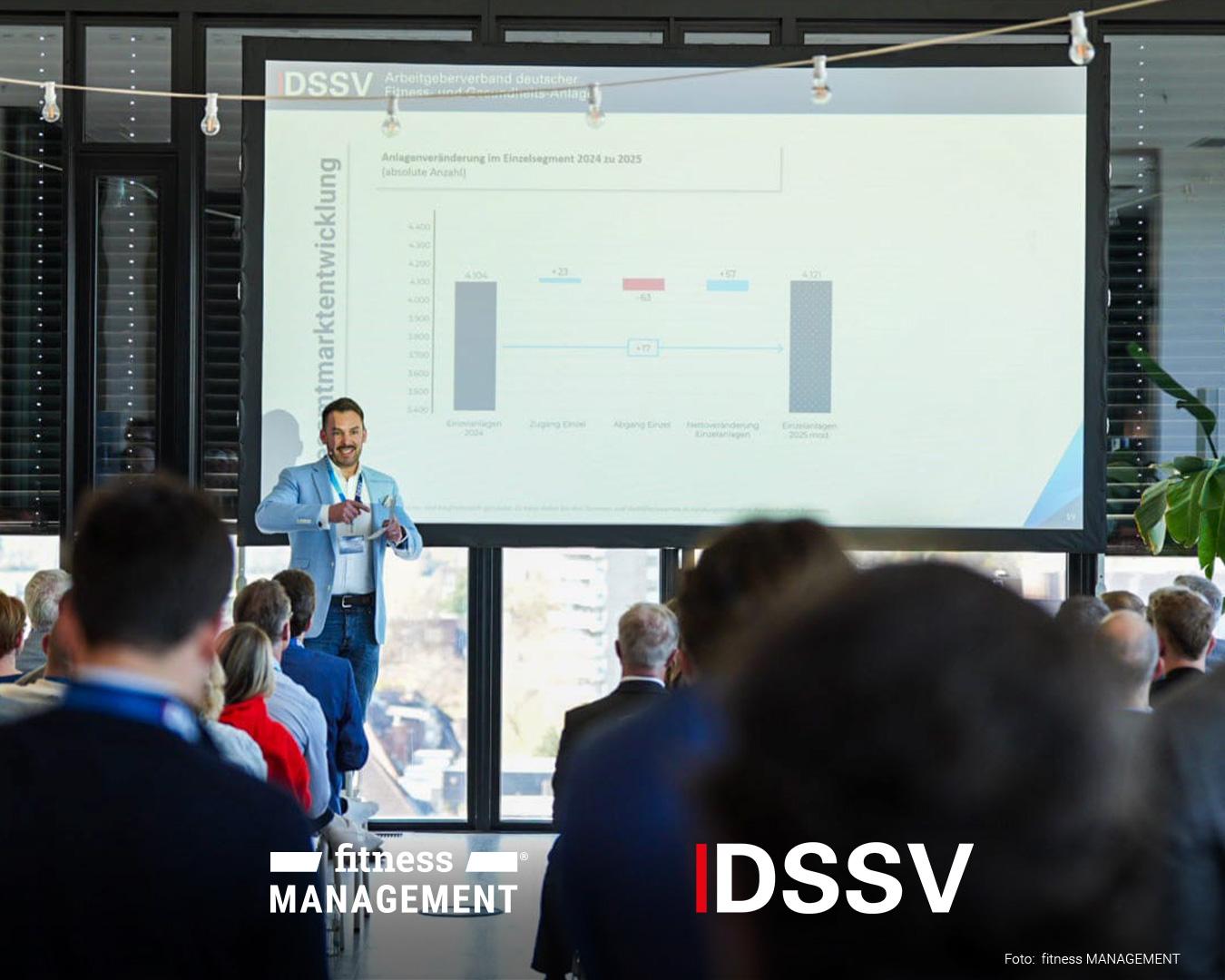 Ein Referent steht vor einer großen Projektionsfläche und zeigt auf ein Diagramm. Im Vordergrund sitzen Zuhörer und blicken in Richtung der Präsentation