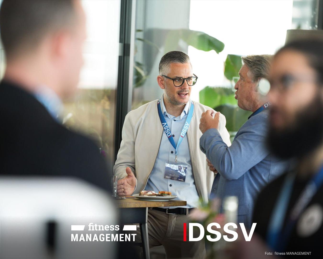 Zwei Männer stehen an einem Stehtisch und unterhalten sich miteinander. Auf dem Tisch steht ein Teller mit Snacks, während im Hintergrund weitere Personen im Gespräch zu sehen sind