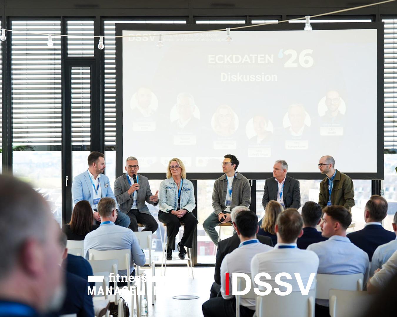 Mehrere Personen sitzen auf einer Bühne bei einer Podiumsdiskussion, während das Publikum ihnen aufmerksam zuhört