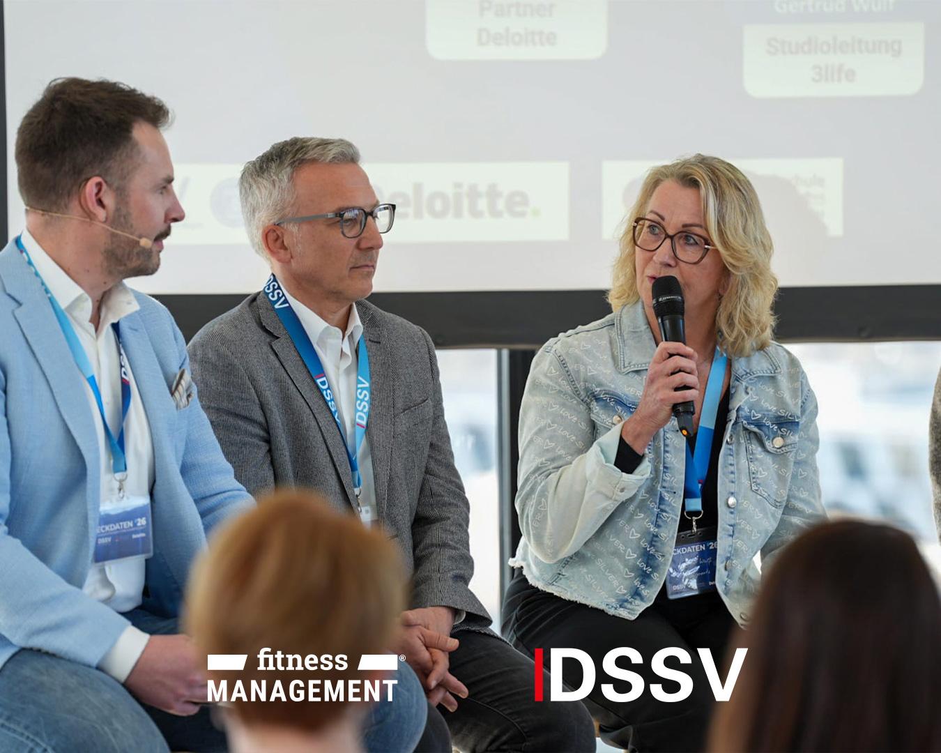 Drei Personen sitzen auf einer Bühne bei einer Podiumsdiskussion, eine Frau spricht in ein Mikrofon, während die anderen zuhören