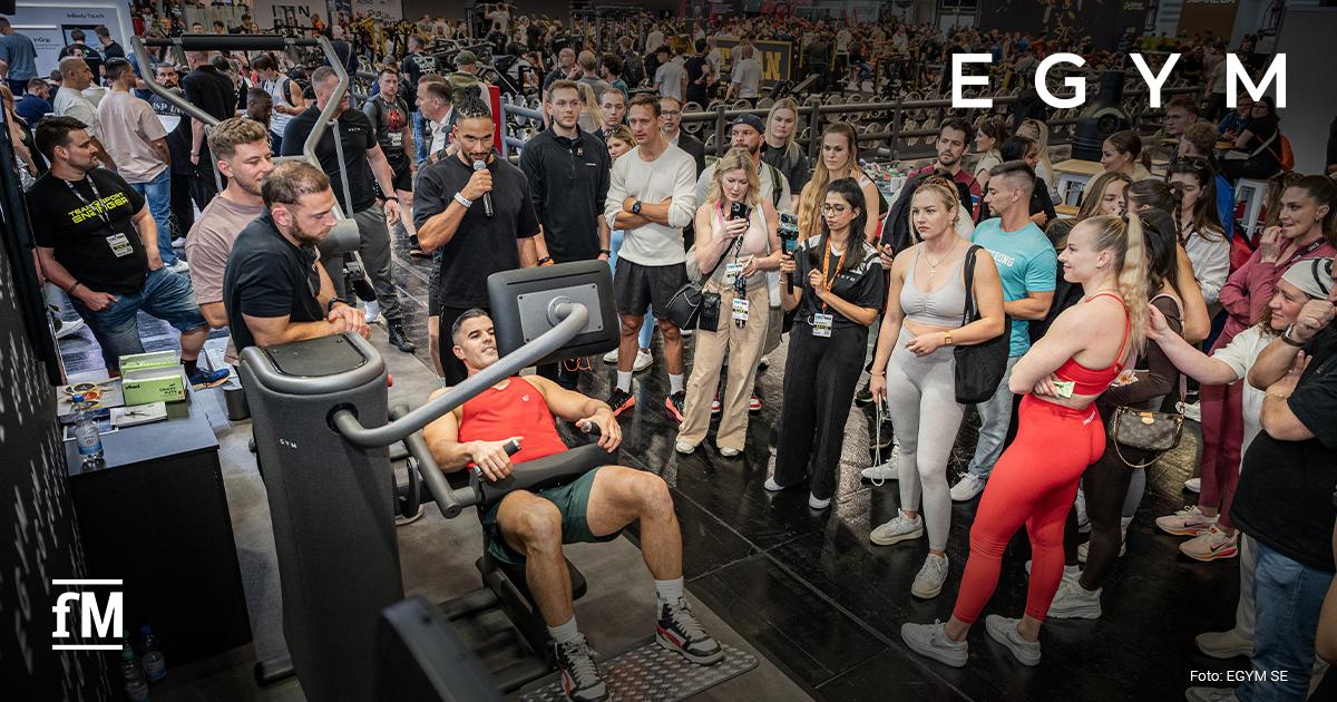 Messehalle mit EGYM-Fitnessgeräten, eine Person trainiert an einer Maschine, viele Menschen stehen um das Gerät herum und beobachten.