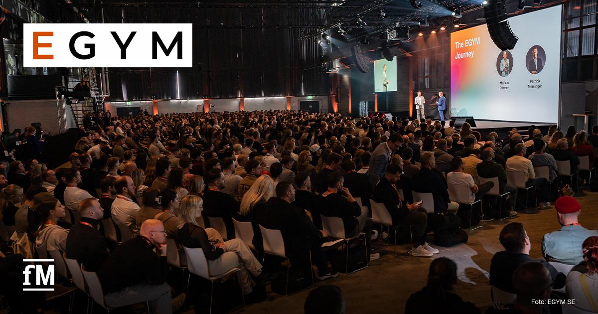 EGYM Kongress 2026 Große Bühne mit Projektionswand „The EGYM Journey“, zwei Sprecher stehen vorn, Publikum sitzt dicht in Reihen, Aufnahme von hinten über Zuschauer hinweg.