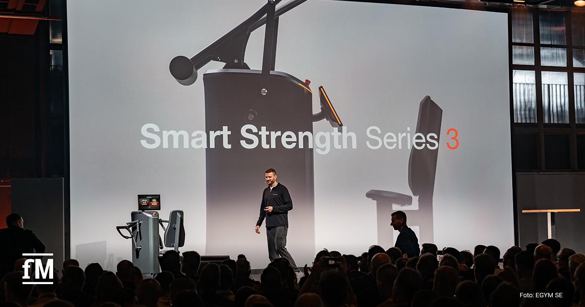 Mann steht neben Trainingsgerät auf Bühne, Hintergrundprojektion zeigt große Gerätegrafik mit Text „Smart Strength Series 3“