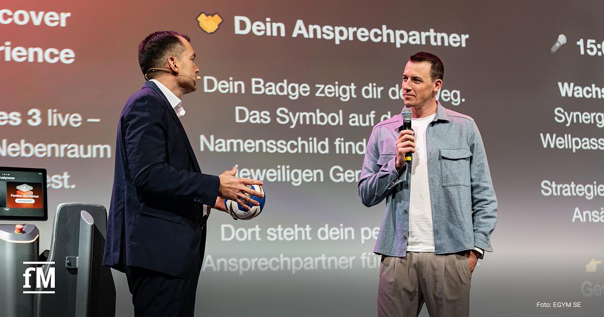 Bühne mit großem Bildschirm im Hintergrund, zwei Personen stehen im Gespräch mit Ball, Publikum sitzt im Vordergrund