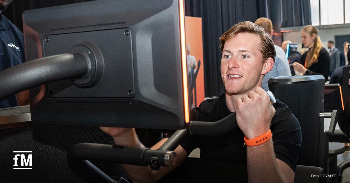 Mann sitzt an Krafttrainingsgerät und bewegt Griffe nach vorn, großes Display am Gerät zeigt Trainingsgrafik, Perspektive nah von vorne