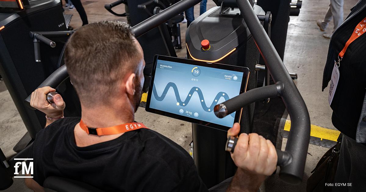 Person sitzt an Trainingsgerät und hält Griffe, Bildschirm zeigt Kurvendiagramm und Trainingsparameter, Kamera über Schulter von hinten