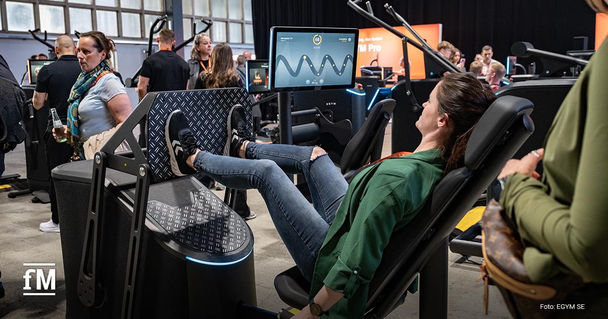 Frau sitzt an Beinpressegerät mit Bildschirm, Füße auf Plattform, andere Besucher stehen im Hintergrund in Ausstellungshalle
