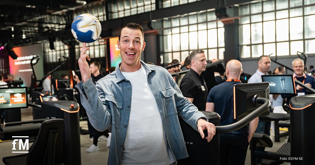 Mann hält Ball und steht zwischen Krafttrainingsgeräten in Messehalle, blickt zur Kamera, Geräte und Besucher im Hintergrund
