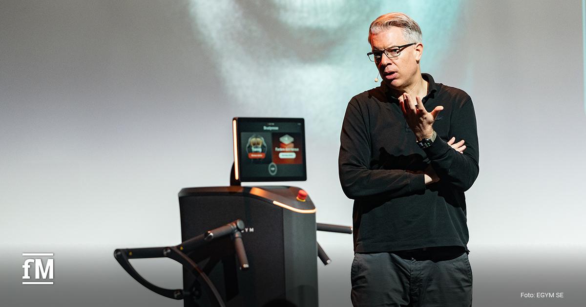 : Sprecher steht neben Trainingsgerät und gestikuliert mit Hand, Gerät mit Bildschirm neben ihm, Hintergrund helle Projektionsfläche