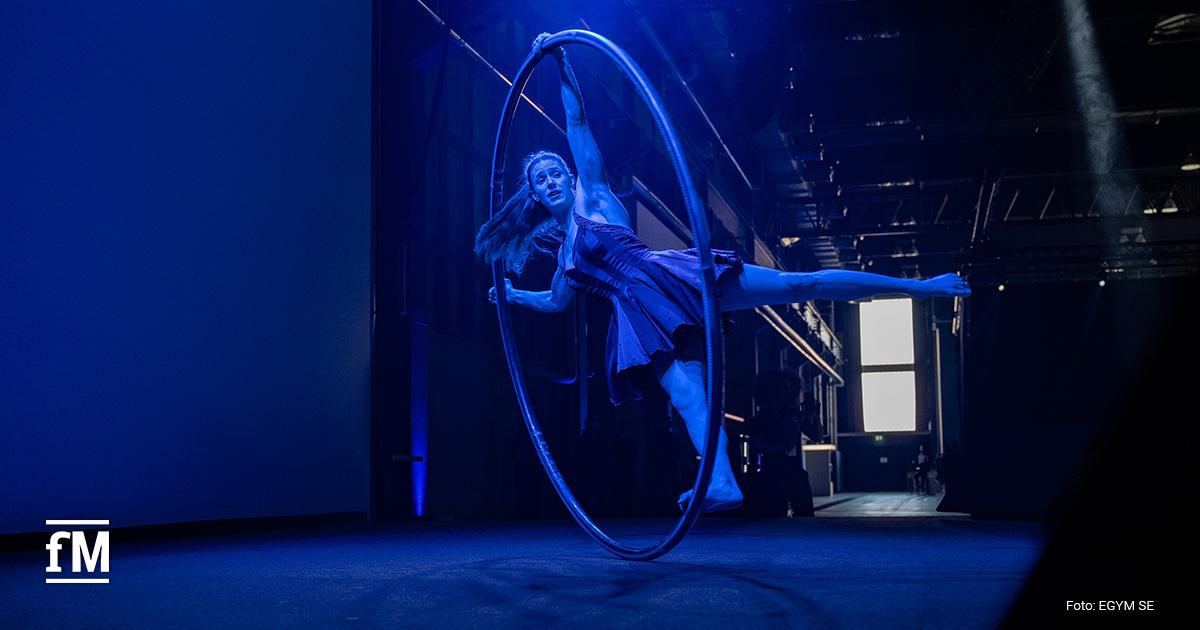 Bühne in dunklem Raum mit blauem Licht, eine Artistin bewegt sich in einem großen Metallring, Hintergrund mit Vorhang und seitlichem Lichtspot, Kamera frontal aus mittlerer Distanz