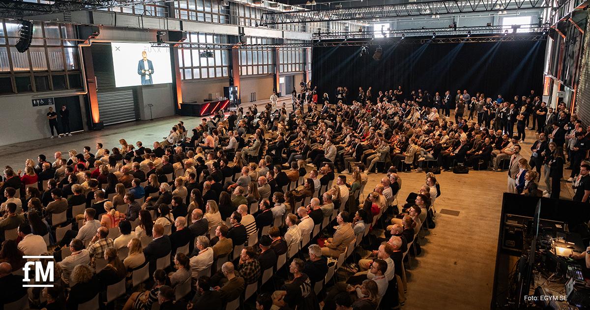 Große Veranstaltungshalle mit dicht besetzten Stuhlreihen, Bühne links mit Präsentationsbildschirm und Sprecher, Blick schräg von oben über Publikum und Technikbereich