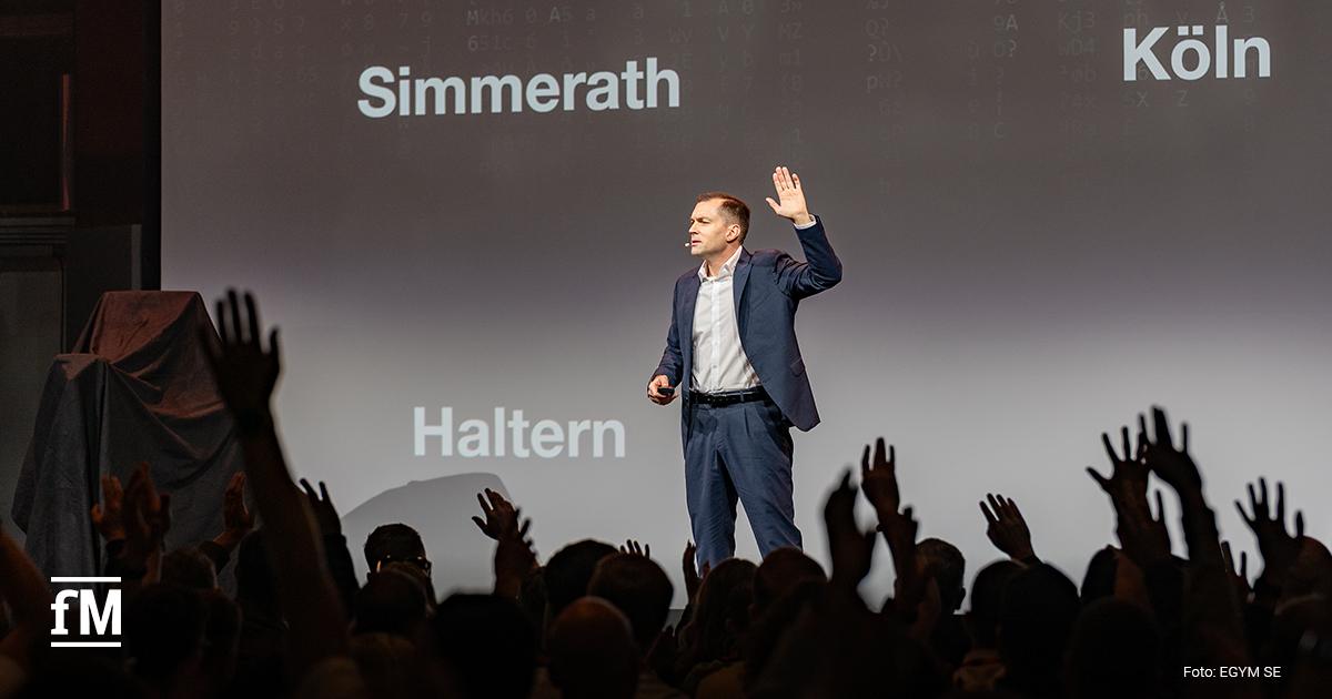 Mann auf Bühne hebt eine Hand, viele Personen im Publikum heben ebenfalls die Hände, Hintergrund mit Projektionswand und Ortsnamen