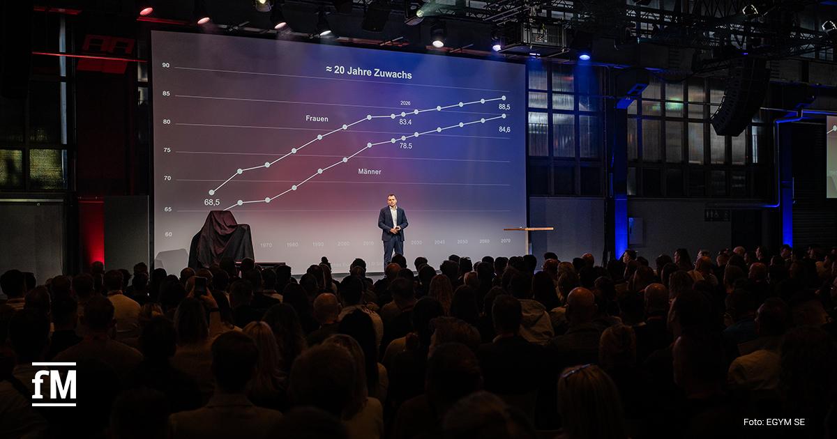 Vortragender steht vor großer Grafikprojektion mit Linienchart zu Wachstum über Jahre, Publikum sitzt im Vordergrund im dunklen Saal.