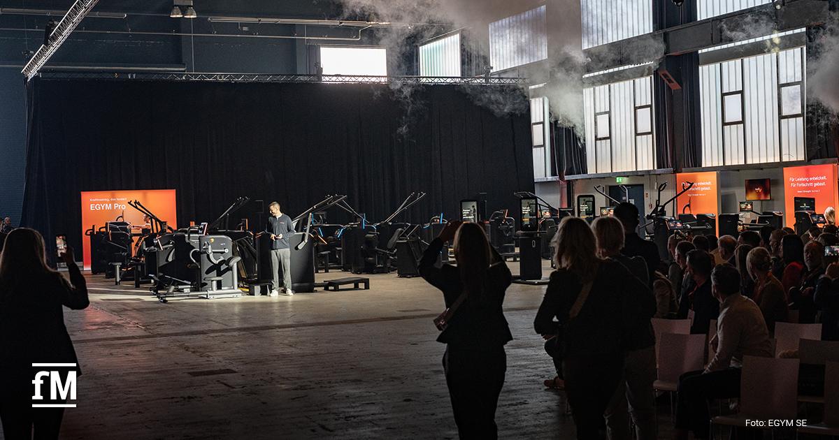 Große offene Halle mit mehreren Krafttrainingsgeräten und digitalen Displays, Besucher stehen und fotografieren Geräte im Vordergrund