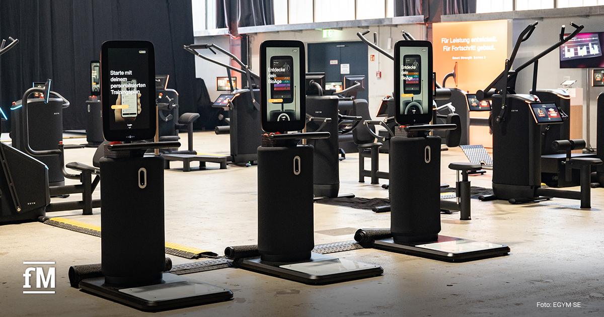 Mehrere schwarze Trainingsgeräte mit großen vertikalen Displays stehen nebeneinander in Messehalle, Fensterfront im Hintergrund