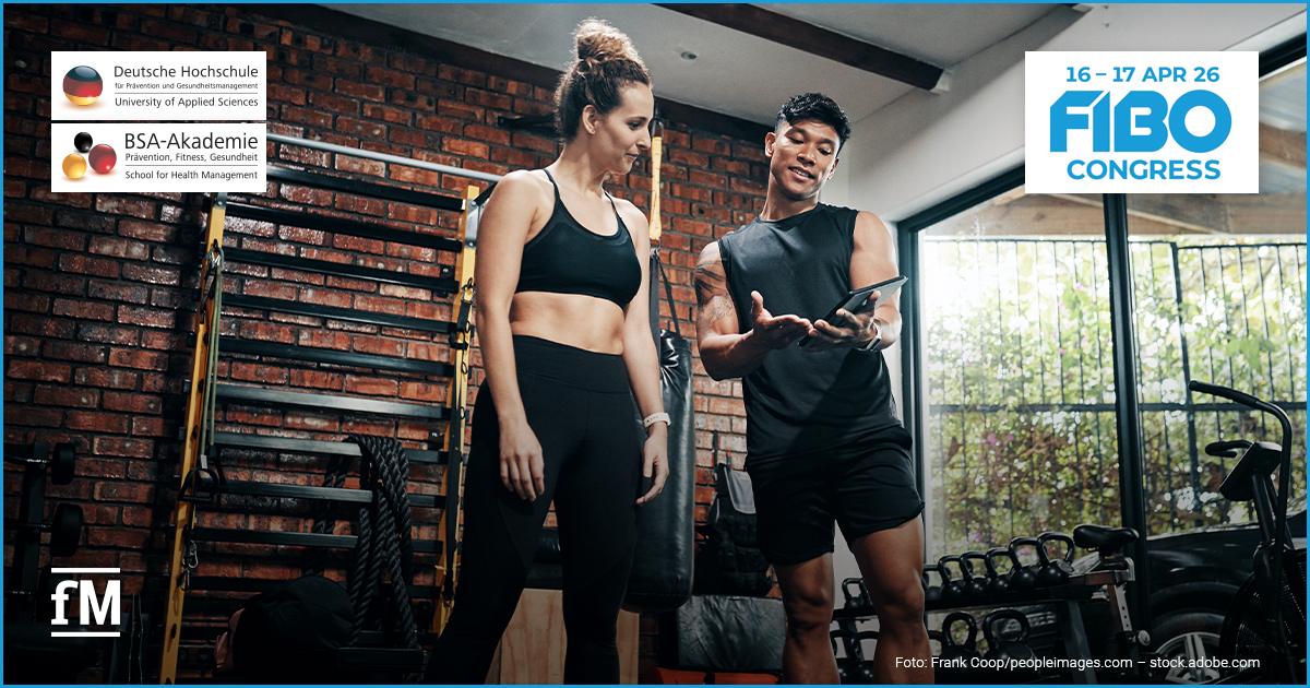 FIBO Congress 2026: Vorträge KI Ein Trainer und eine Sportlerin stehen in einem Fitnessstudio vor einer Backsteinwand. Der Trainer hält ein Tablet in der Hand und erklärt etwas, während im Hintergrund Trainingsgeräte wie Kettlebells und Seile zu sehen sind