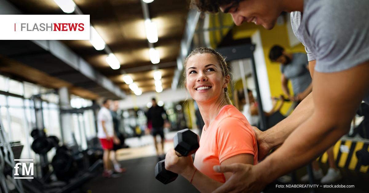 Eine lächelnde Frau im Fitnessstudio hält eine Kurzhantel, während ein Trainer ihr beim Schultertraining hilft; im Hintergrund sind weitere Sportgeräte und trainierende Personen unscharf zu sehen.