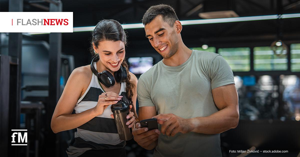 Fitness Flash News #04/2026 Zwei junge Erwachsene im Fitnessstudio lächeln und schauen gemeinsam auf ein Smartphone; die Frau trägt Kopfhörer um den Hals und hält einen Shaker. Oben links steht „FLASH NEWS“.